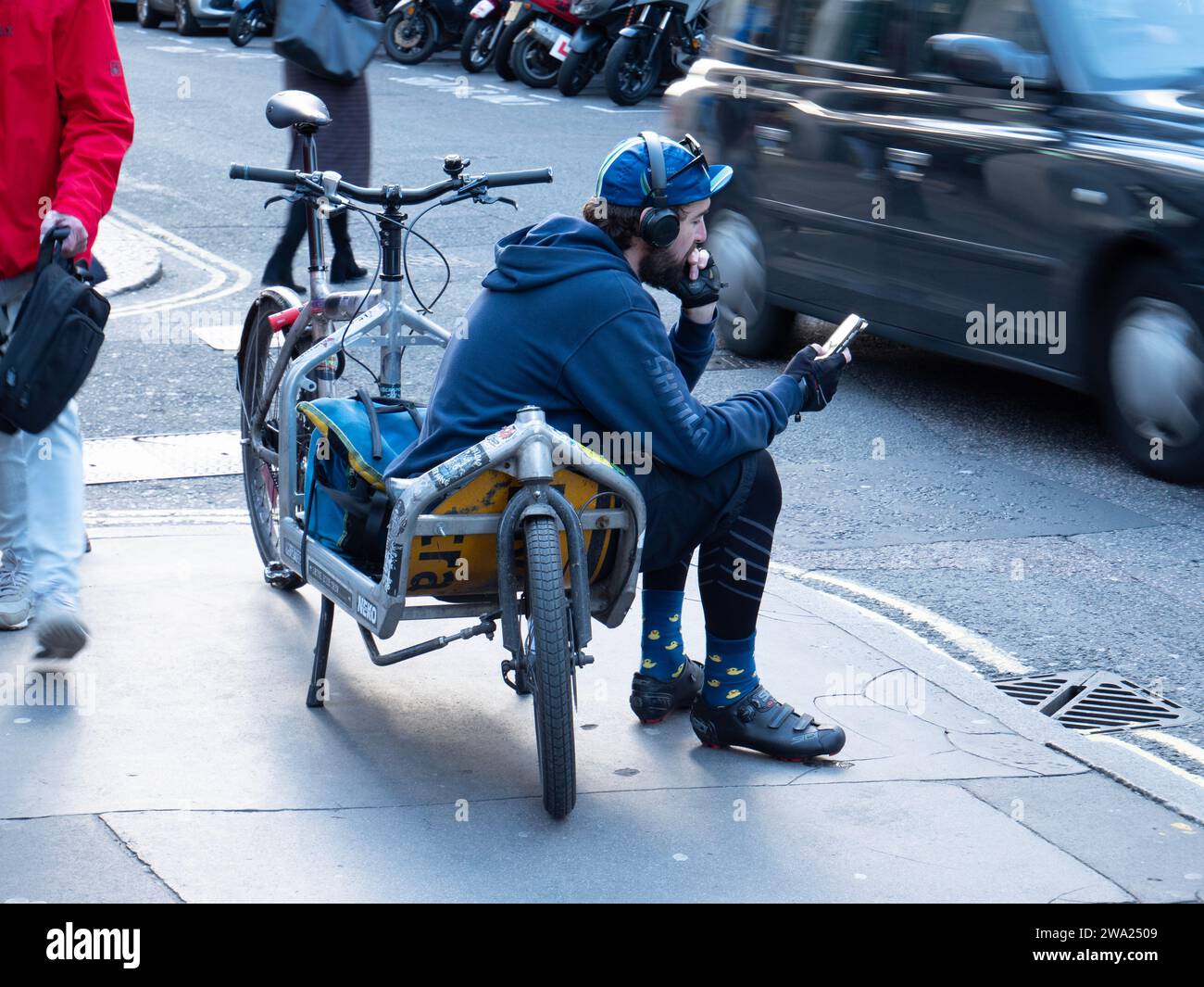 Londres, Royaume-Uni , vélo Courier, cargo vélo messager en attente d'emplois à Soho, Londres, regardant le téléphone portable assis sur le vélo dans la rue Banque D'Images