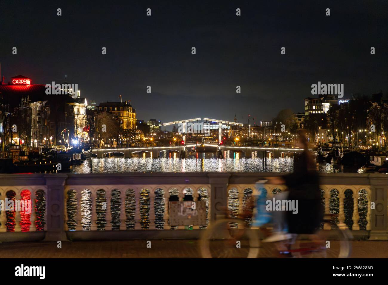 Vue nocturne d'Amsterdam avec le Skinny Bridge (Magere Brug), théâtre carré, avec mouvement flou cycliste passant au premier plan Banque D'Images
