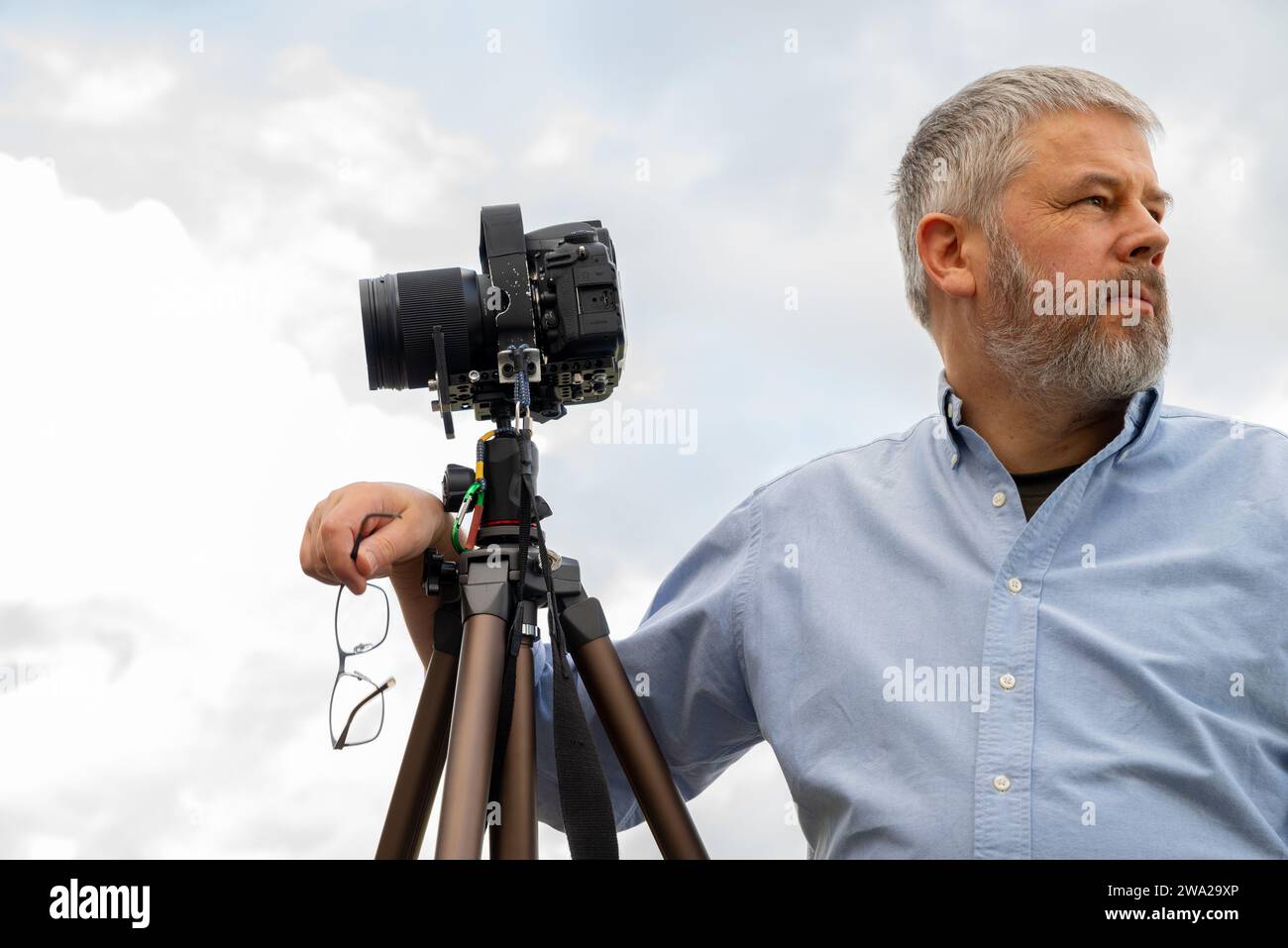 Photographe au travail, cheveux gris et barbe complète, 56 ans, chemise légère, lunettes mises en main, appareil photo reflex 35mm sur trépied, ciel couvert gris Banque D'Images