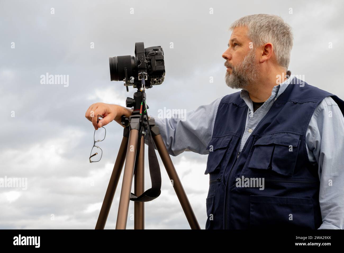 Photographe au travail, cheveux gris et barbe complète, 56 ans, gilet photo bleu foncé, lunettes à la main, appareil photo reflex 35mm sur trépied, ciel couvert gris Banque D'Images