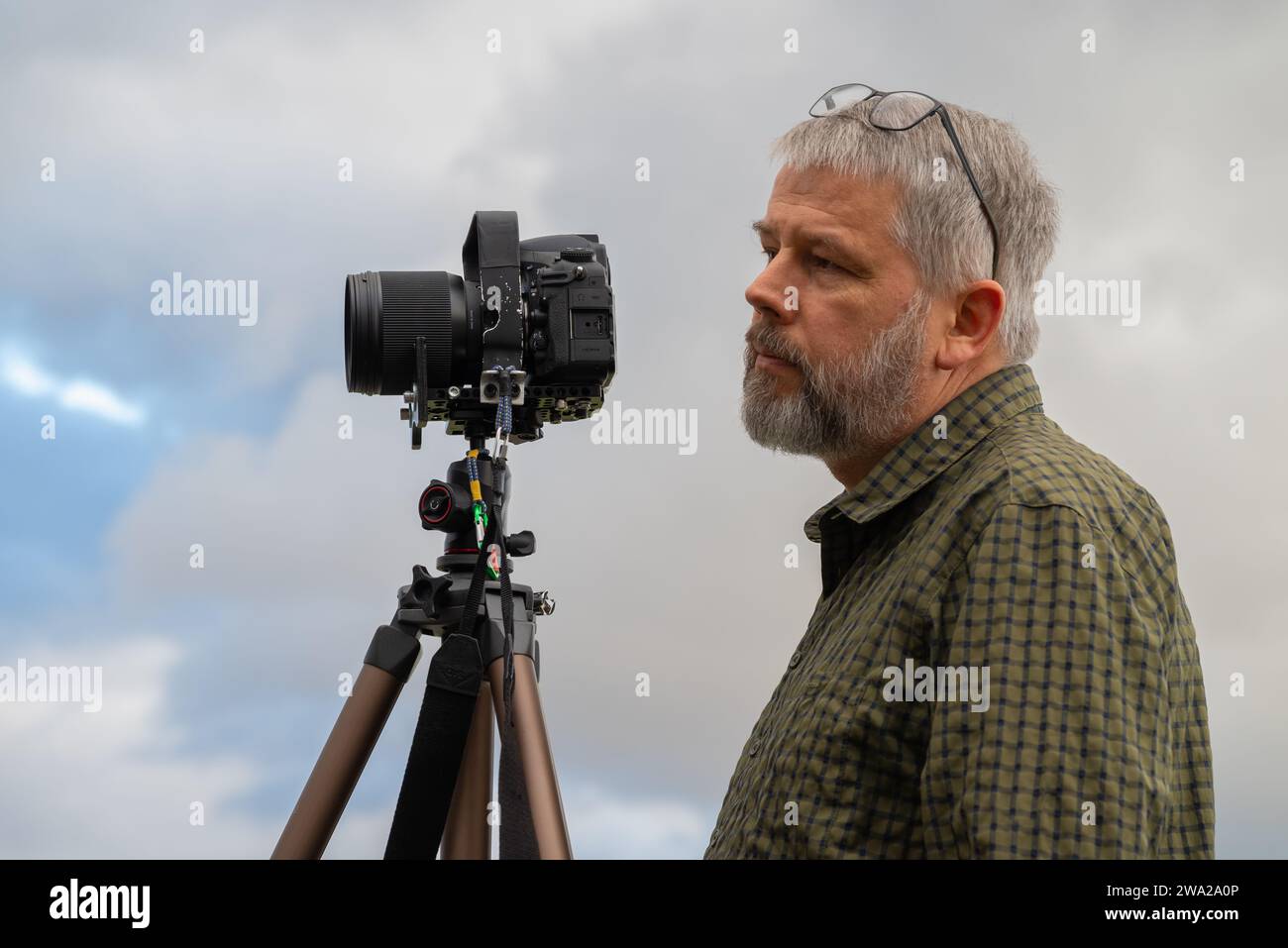 Photographe au travail, cheveux gris et barbe complète, 56 ans, regardant devant l'appareil vers le sujet, lunettes poussées, chemise à carreaux vert olive, Banque D'Images