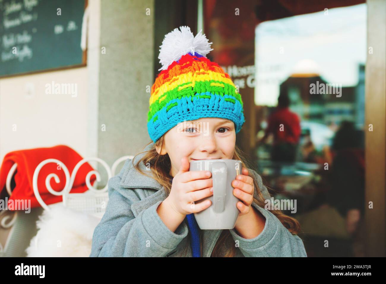 Adorable petite fille de 6 ans buvant du chocolat chaud dans un café d'hiver, tenant une grande tasse, portant un manteau gris et un chapeau coloré Banque D'Images