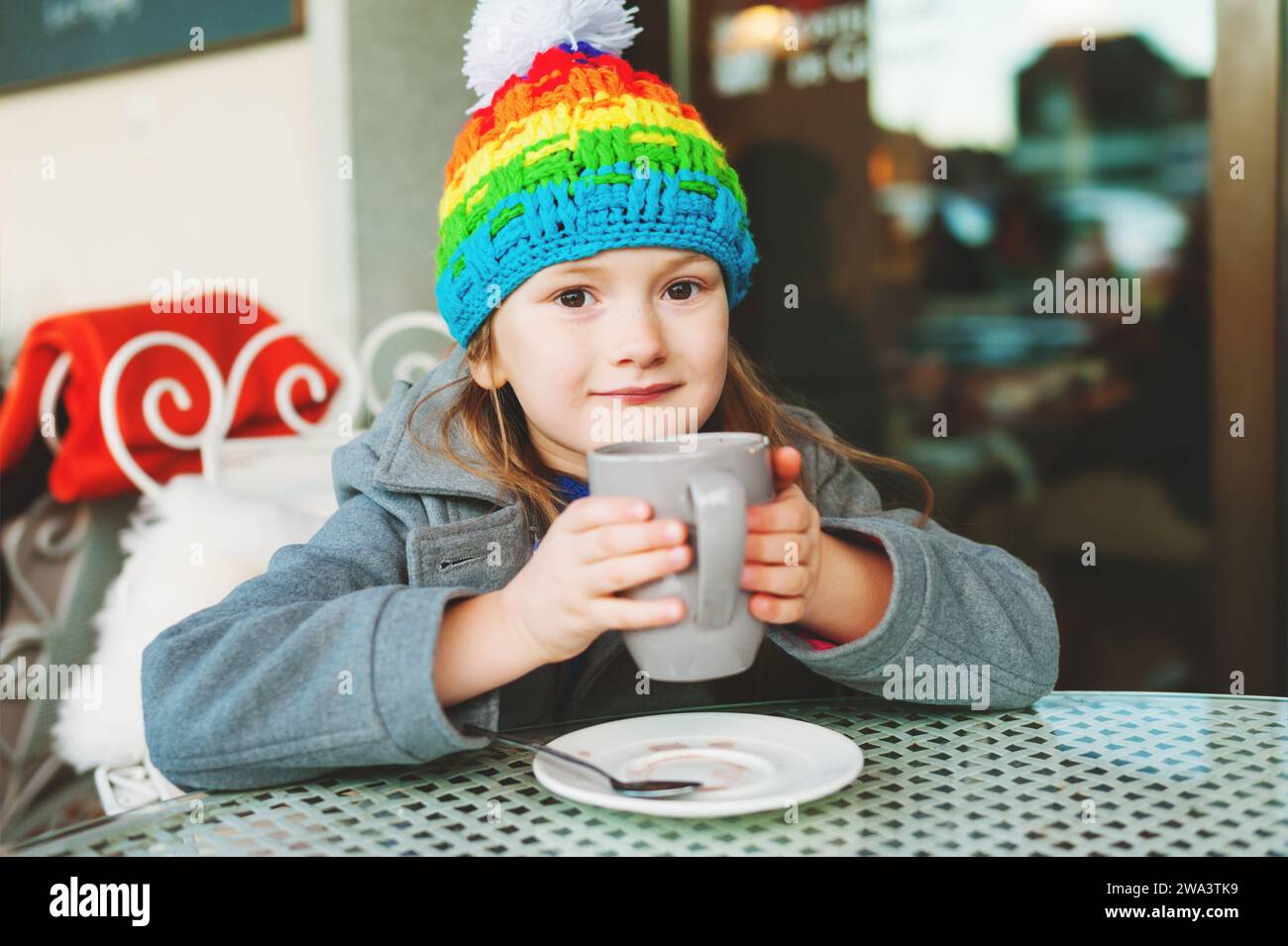 Adorable petite fille de 6 ans buvant du chocolat chaud dans un café d'hiver, tenant une grande tasse, portant un manteau gris et un chapeau coloré Banque D'Images