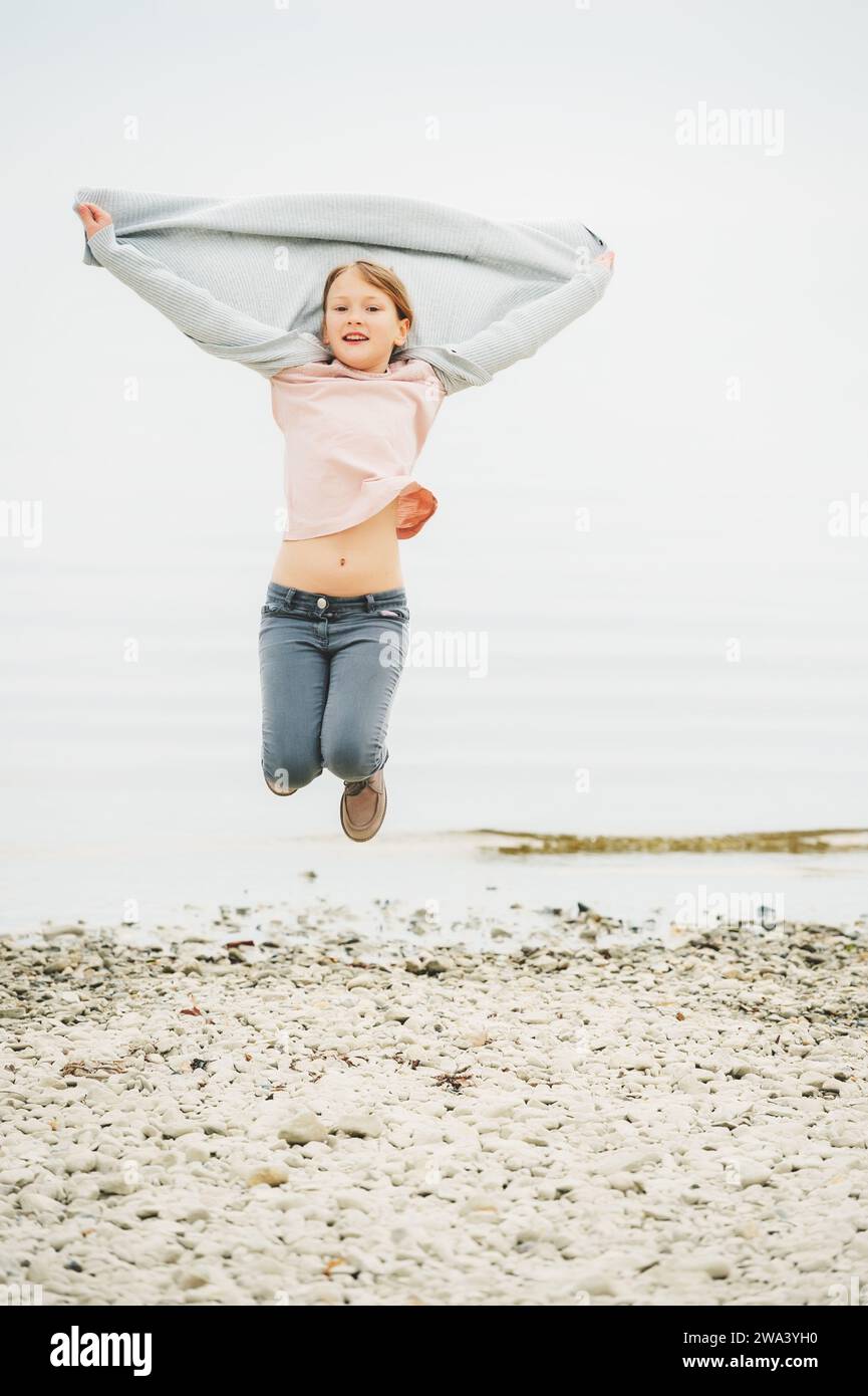 Adorable petite fille de 8-9 ans jouant au bord du lac, portant un pantalon gris et un long cardigan, sautant en l'air Banque D'Images