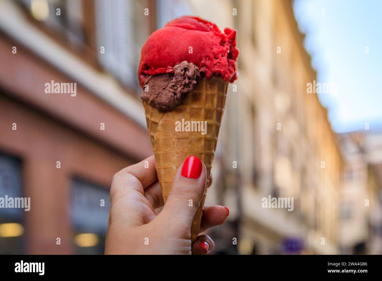 Main de femme tenant le chocolat artisanal et le gelato framboise dans un cône, avec des colombages dans le carré d ou quartier de Strasbourg, Alsace, France Banque D'Images
