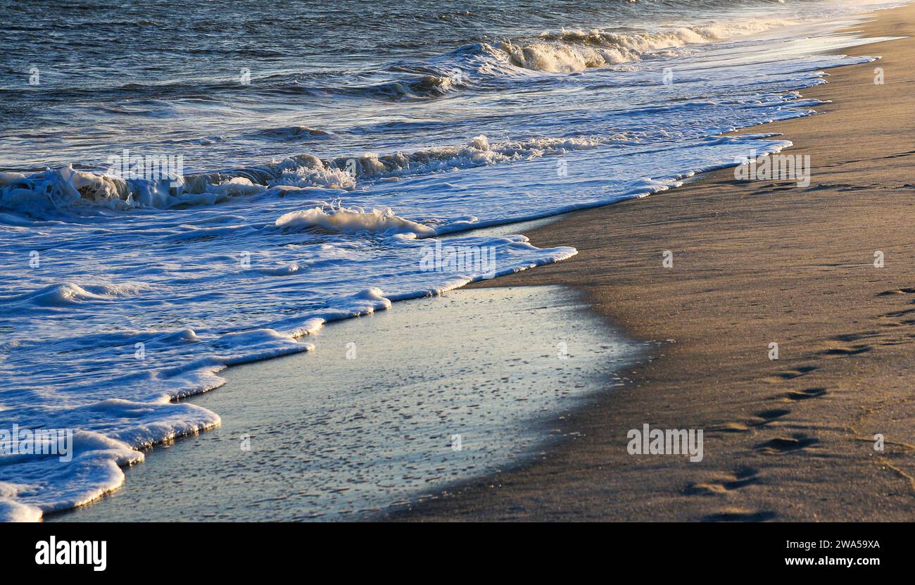 Une plage de sable avec des vagues arrivant sur le rivage et la lumière du soleil couchant se reflétant sur l'eau. Banque D'Images