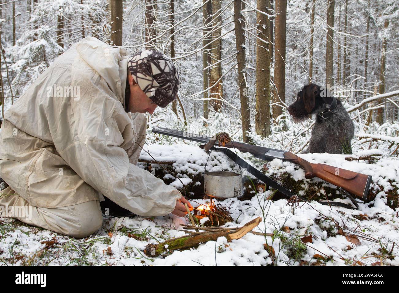 un chasseur vêtu de camouflage blanc tente de construire un feu dans la forêt d'hiver en utilisant des allumettes, et son chien est assis à côté de lui Banque D'Images
