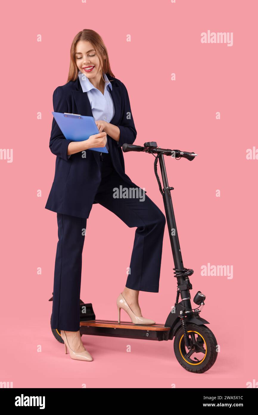 Portrait de jeune femme d'affaires avec presse-papiers et kick scooter sur fond rose Banque D'Images