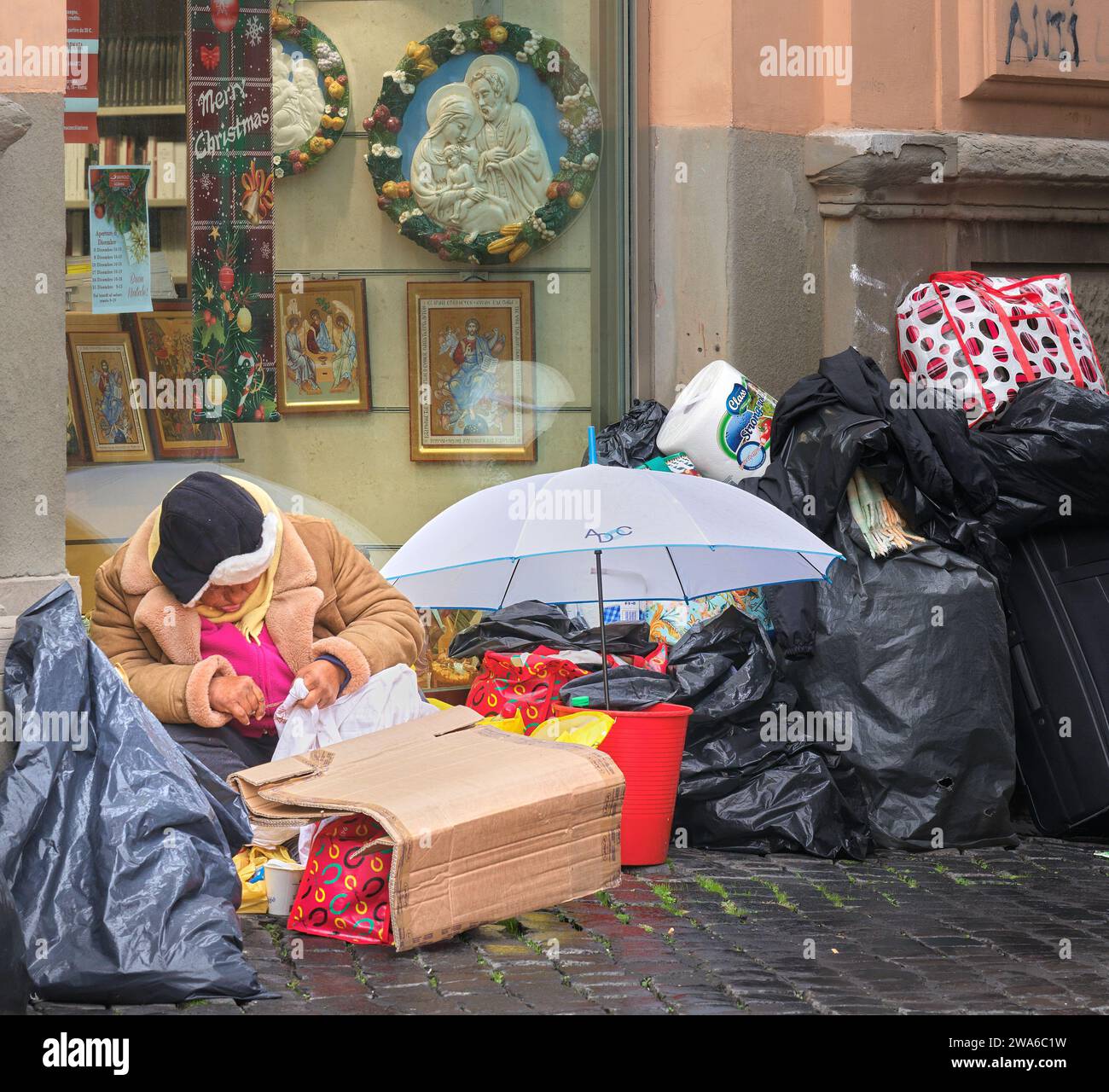 En bas et dehors à Rome ; une personne sans abri est assise avec ses possessions dans des sacs en plastique devant un magasin avec «Joyeux Noël», dans une rue de la cité du Vatican. Banque D'Images