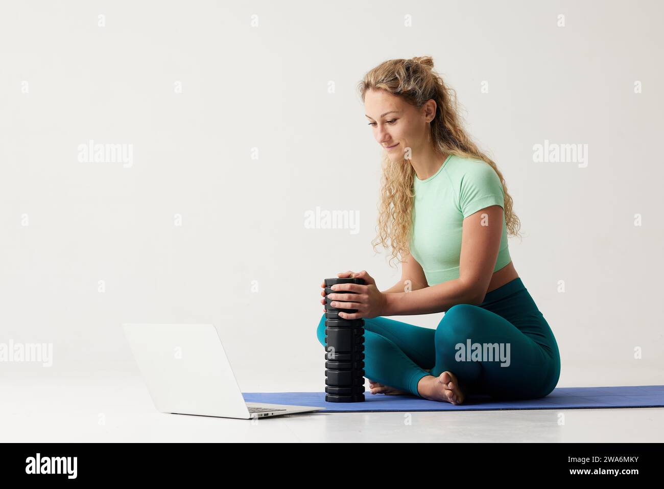 fille tenant un rouleau de massage tout en étant assise sur le sol et près d'un ordinateur portable, rouleau de massage pour la remise en forme et le yoga, entraînement en ligne à la maison Banque D'Images