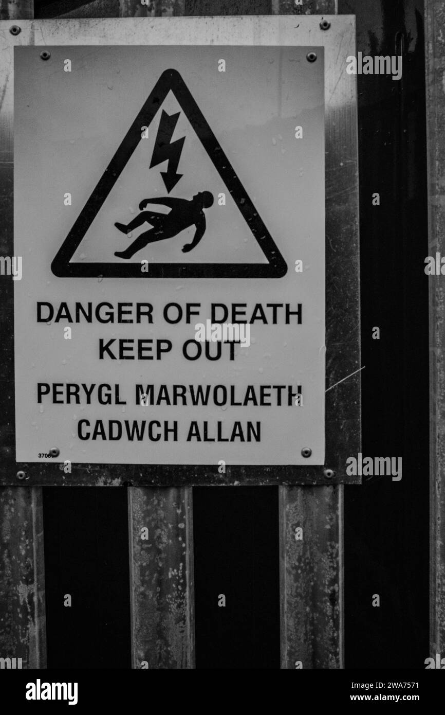 Panneau d'avertissement de danger de mort monté sur une clôture de sécurité de transformateur d'électricité. Le libellé est en anglais et en gallois. B&W. Banque D'Images