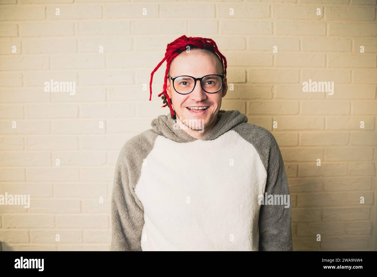 jeune homme hipster attrayant avec des dreadlocks rouges dans des lunettes et un pull blanc sur le fond d'un mur de briques. Banque D'Images