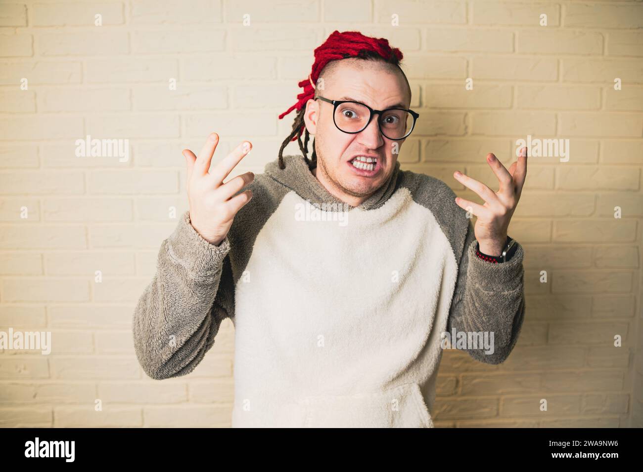 Un bel homme adulte avec des lunettes et des dreadlocks rouges est en colère contre un mur de briques blanches. Banque D'Images