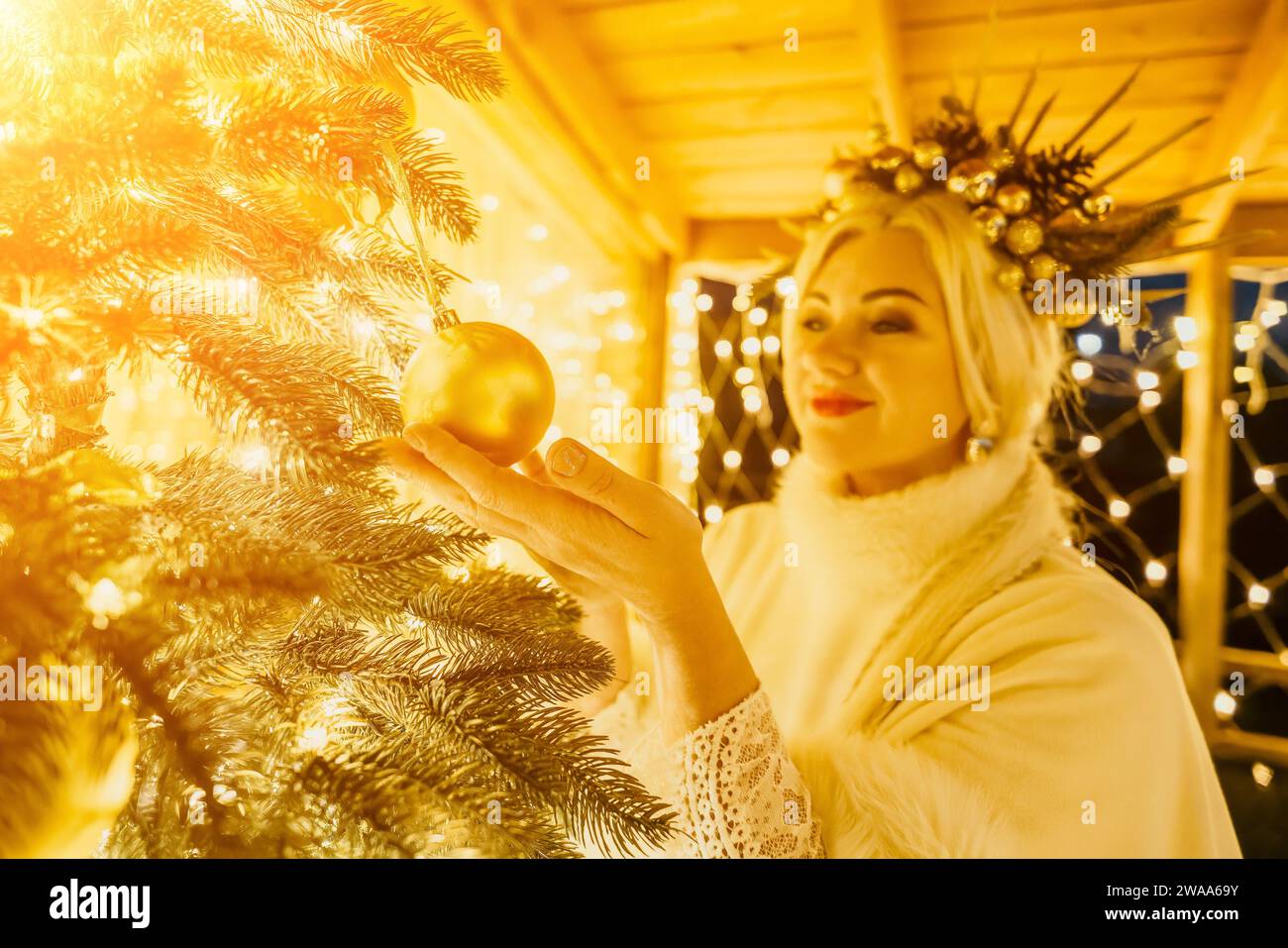 Une femme blonde en robe blanche et une couronne d'ornements en or décorent l'arbre de Noël avec des ornements en or et des lumières. L'arbre est décoré d'or Banque D'Images