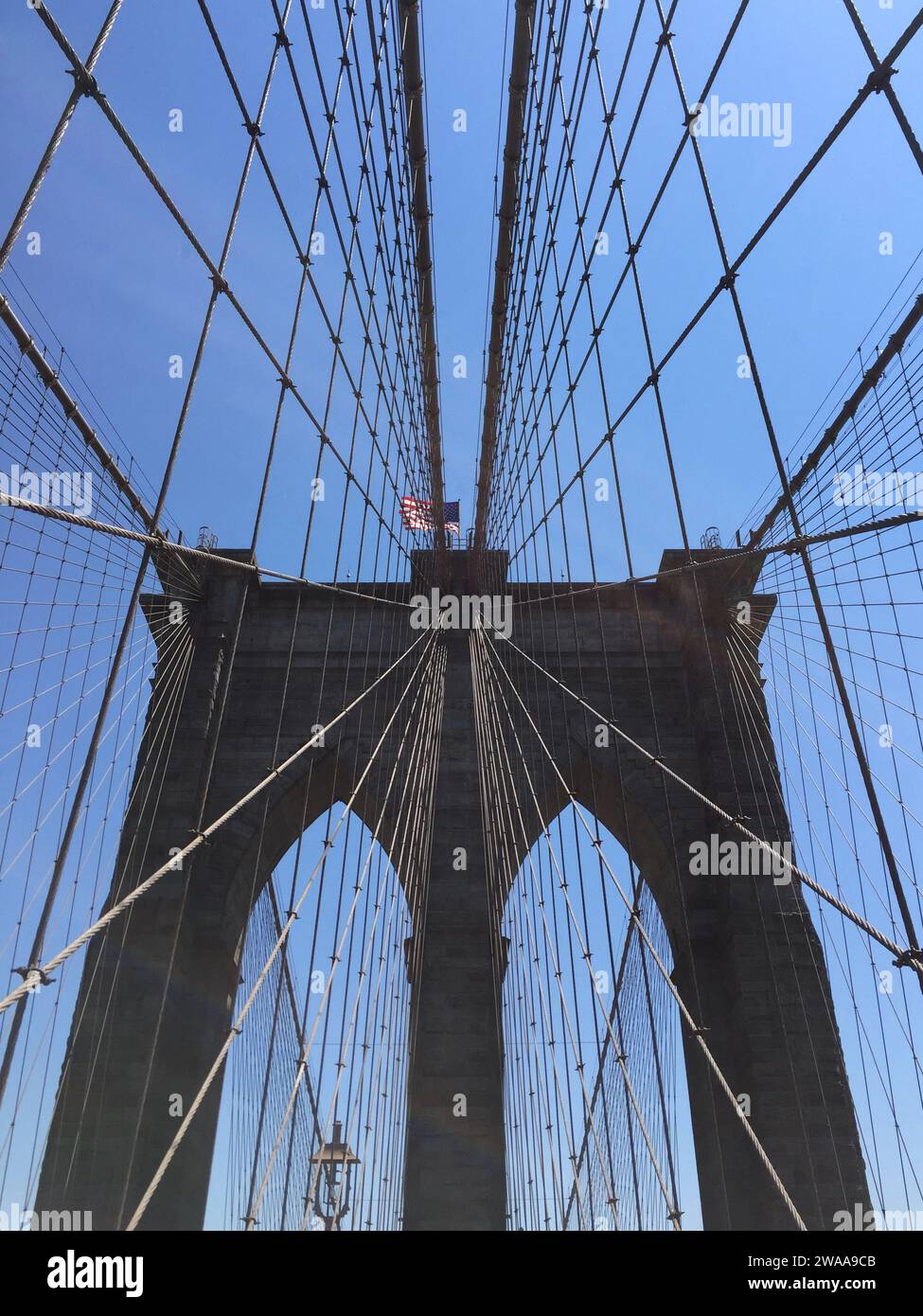 Le pont de Brooklyn s'étend au-dessus, ses câbles emblématiques formant un motif frappant contre le ciel bleu, couronné par le drapeau américain flottant. Banque D'Images