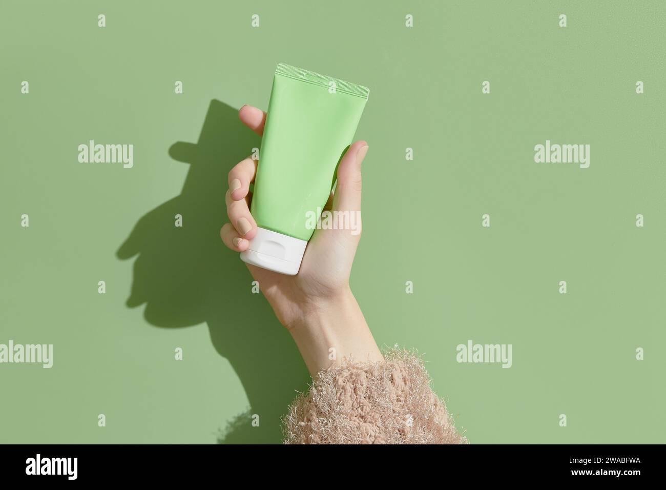 Une main de femme dans un pull tient un tube de crème sur un fond vert. Soins de beauté auto-soignés Banque D'Images