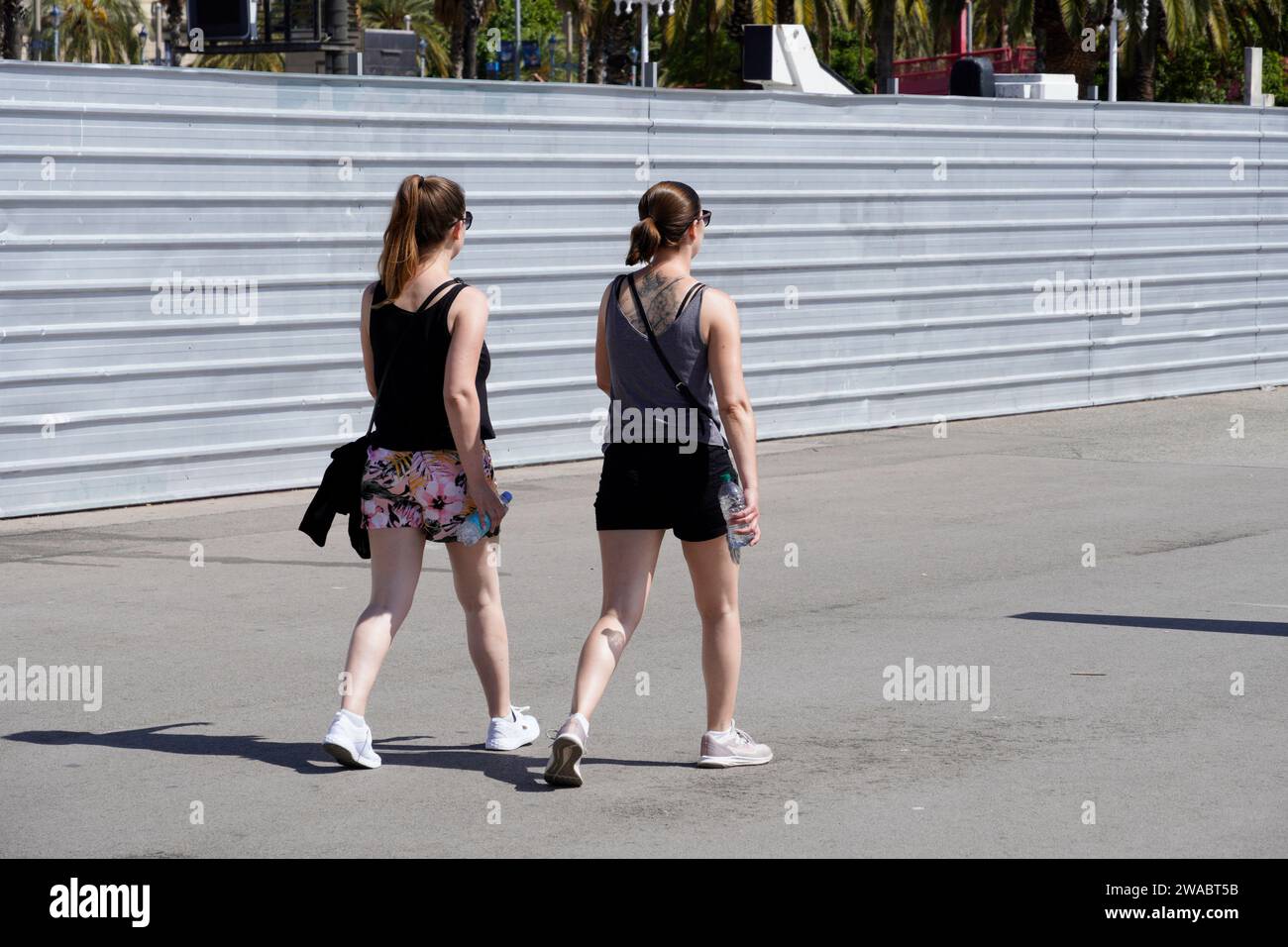 Barcelone, Espagne - 26 mai 2022 : deux jeunes femmes avec leurs cheveux attachés et des lunettes de soleil marchent ensemble avec des bouteilles d'eau dans leurs mains à travers t Banque D'Images