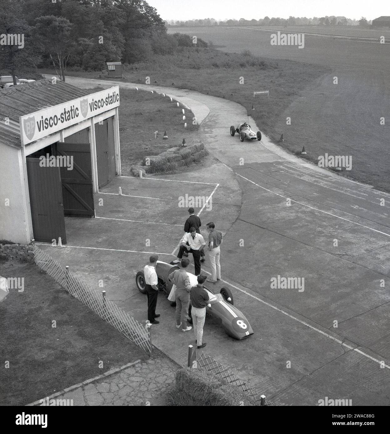 1962, historique, vue aérienne de l'activité aux écuries de course automobile de Geoff Clarke, à l'extérieur de l'aérodrome de Finmere, Buckinghamshire, Angleterre, Royaume-Uni. RAF Finmere était une base de bombardiers de la Seconde Guerre mondiale qui, avec trois pistes, avait une grande route périphérique et c'était celle-ci qui a été utilisée pour le circuit de l'école automobile, qui a vu des élèves conduire des voitures de course Cooper, avec une sur la piste voir sur la photo. Banque D'Images