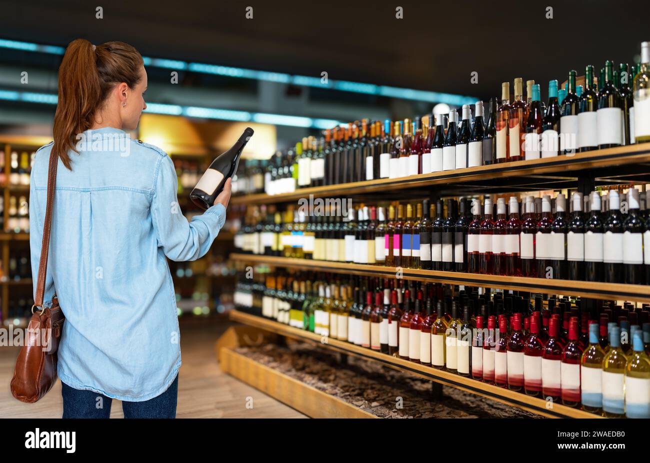 Vue arrière de la cliente femme se tient devant les étagères avec des bouteilles de vin choisissant le vin dans le magasin d'alcool. Banque D'Images