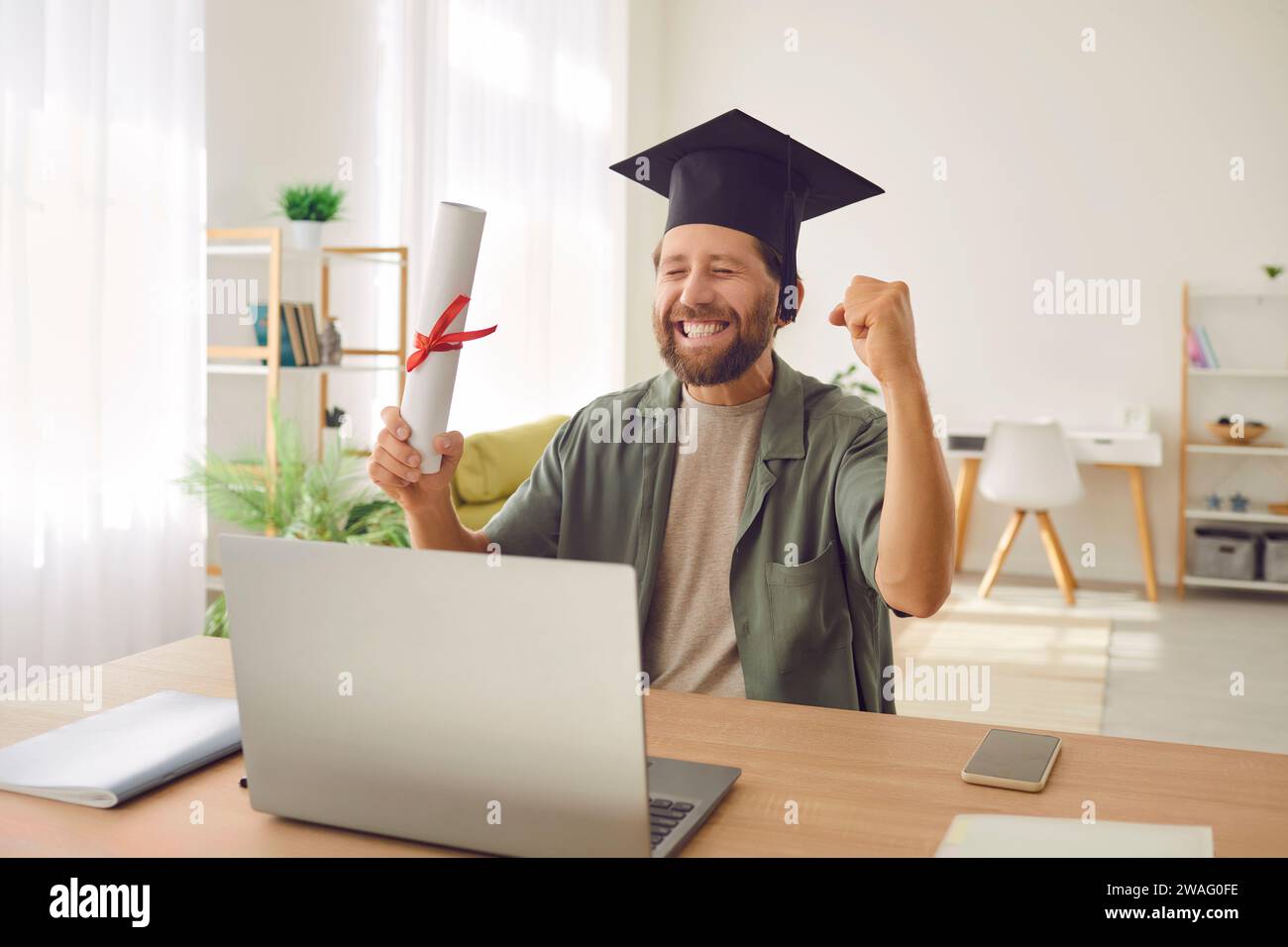 Homme diplômé assis au bureau avec diplôme faisant le geste oui pendant l'appel vidéo en ligne. Banque D'Images