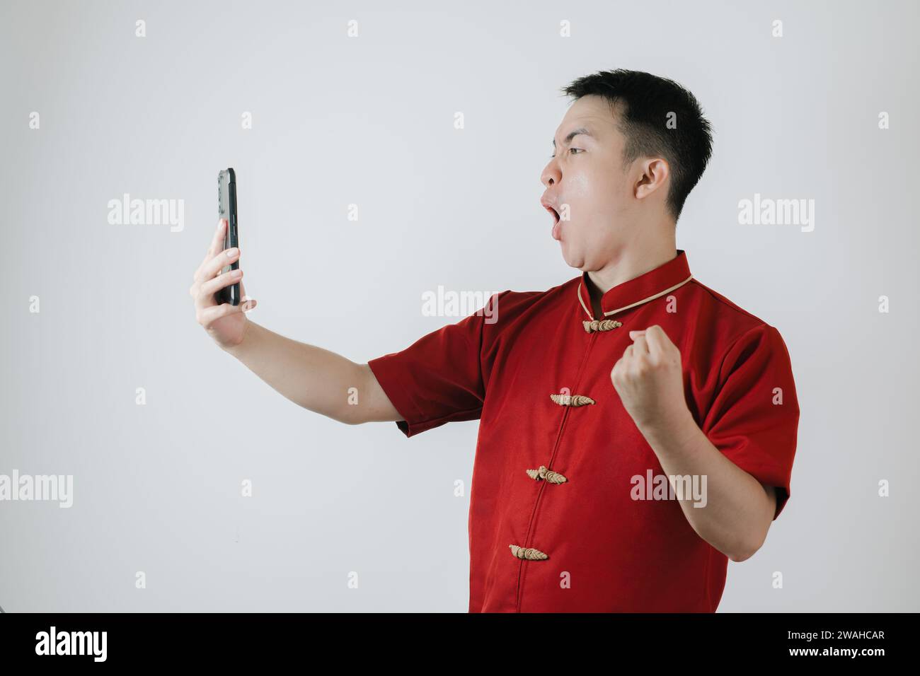 Visage heureux de l'homme asiatique portant le tissu traditionnel chinois ou Cheongsam tout en regardant son smartphone sur fond blanc Banque D'Images