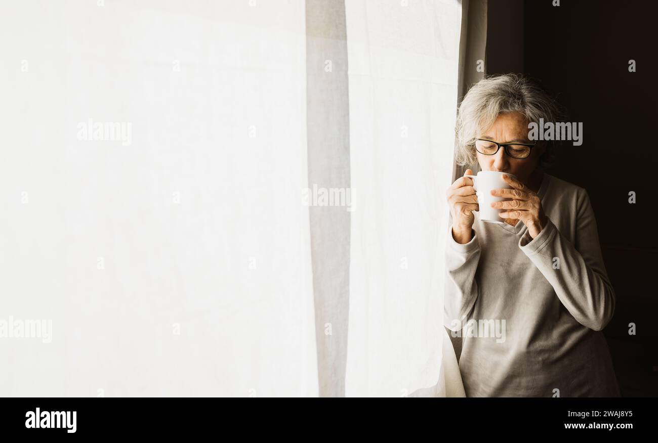 Femme âgée debout près d'une fenêtre et buvant du café sur une journée ensoleillée à la maison. Banque D'Images