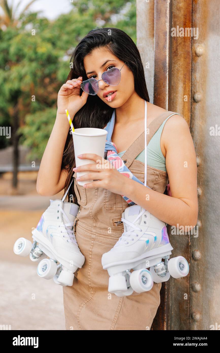 Confiante jeune femme à cheveux longs en tenue décontractée et lunettes debout avec tasse de boisson et patins à roulettes quad blanc sur terrain de sport pavé en pa Banque D'Images