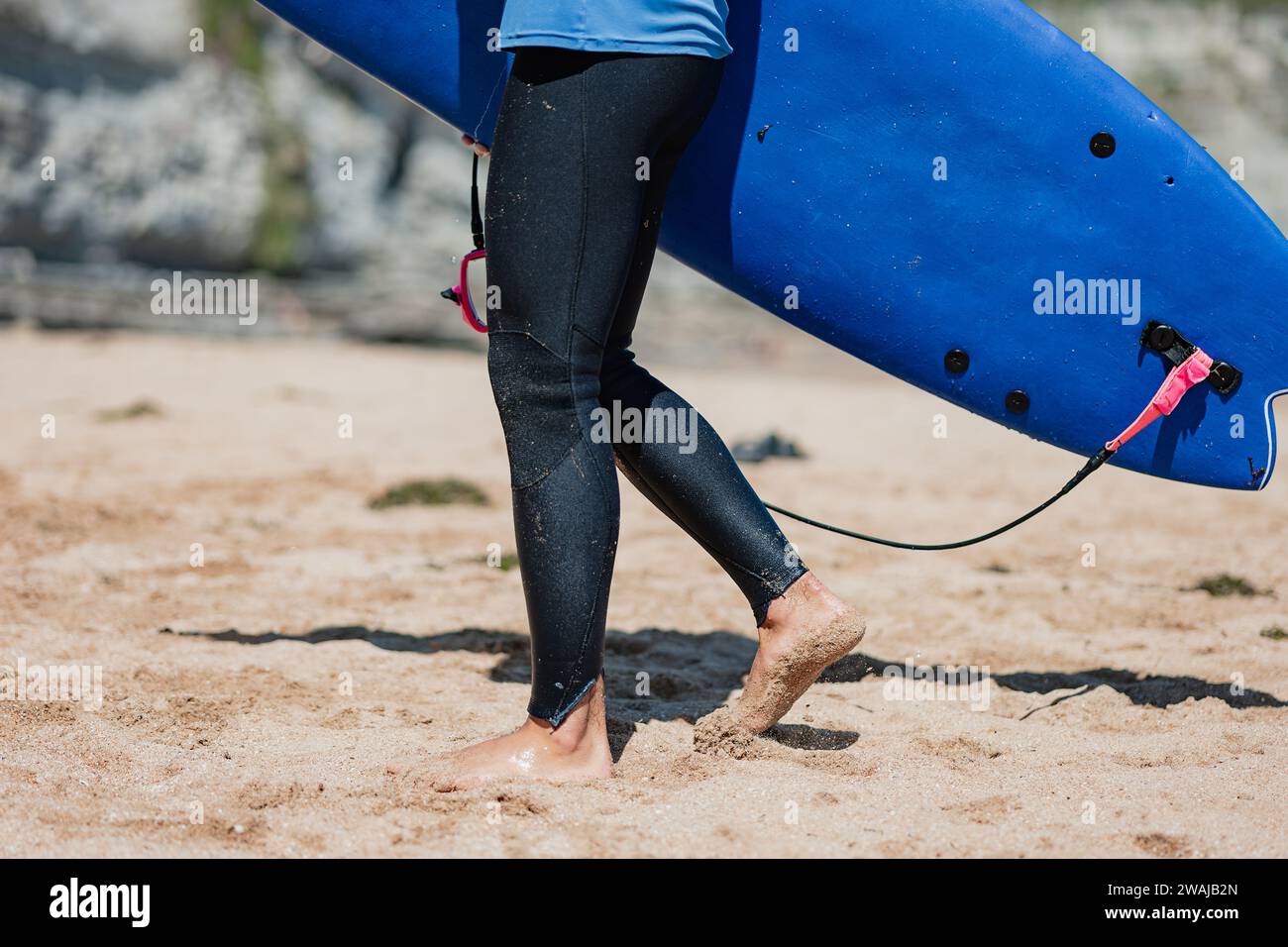 Gros plan du bas du corps d'un surfeur, tenant une planche de surf bleue avec une laisse rose sur une plage de sable Banque D'Images