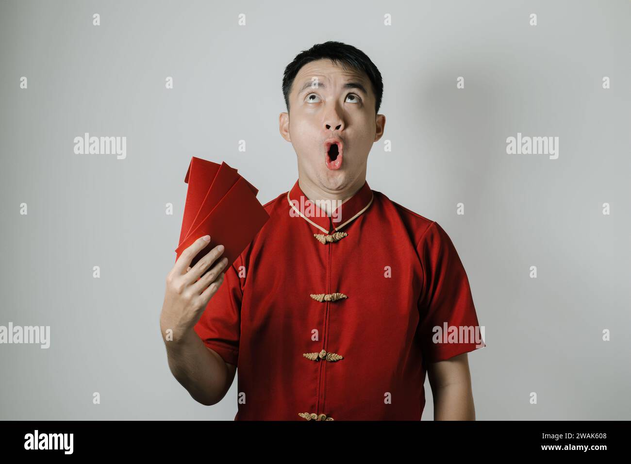 Visage choqué de l'homme asiatique portant Cheongsam ou tissu traditionnel chinois tout en attisant son auto en utilisant angpao ou cadeau monétaire rouge sur fond blanc Banque D'Images