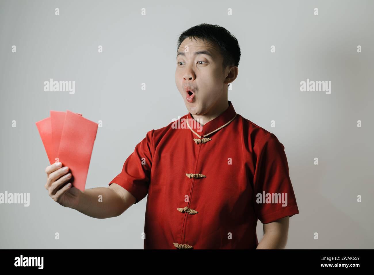 Visage choqué de l'homme asiatique portant Cheongsam ou tissu traditionnel chinois tout en attisant son auto en utilisant angpao ou cadeau monétaire rouge sur fond blanc Banque D'Images