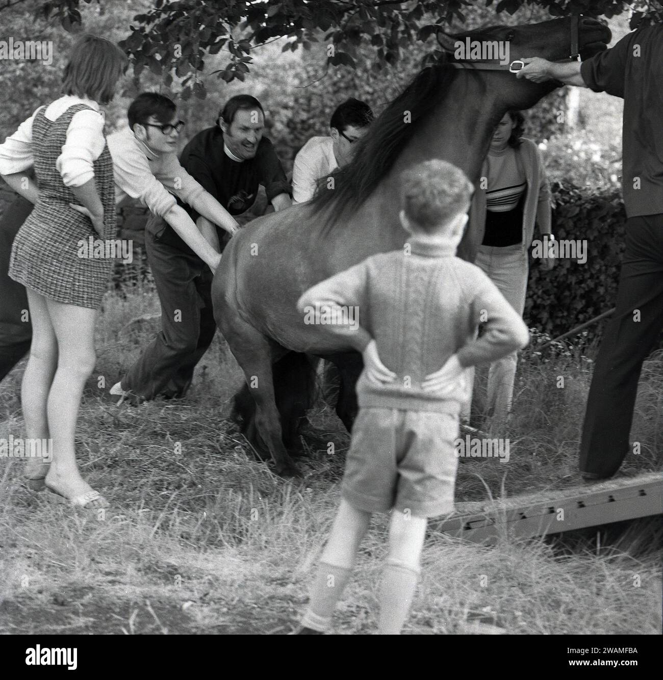 Années 1970, historique, estivale et un garçon et une femme debout dans un champ regardant un certain nombre d'hommes adultes, y compris un vicaire, alors qu'ils tentent de pousser un cheval dans sa remorque ou sa boîte à chevaux à l'extérieur à une foire de vapeur, Angleterre, Royaume-Uni. Banque D'Images