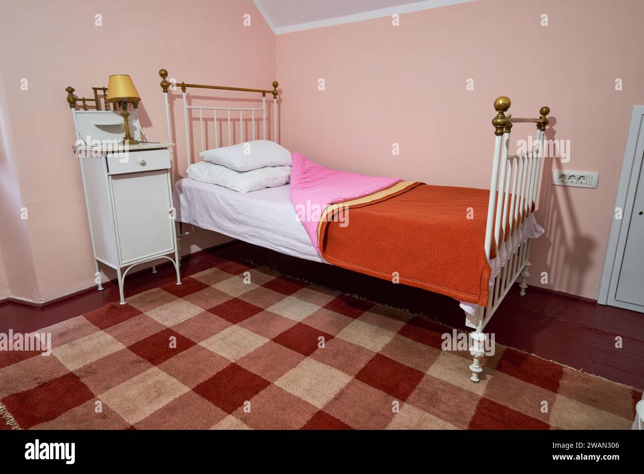 Chambre de type cabine ancienne avec lit en fer blanc, couvre-lits rustiques roses et orange et tapis à carreaux. Banque D'Images