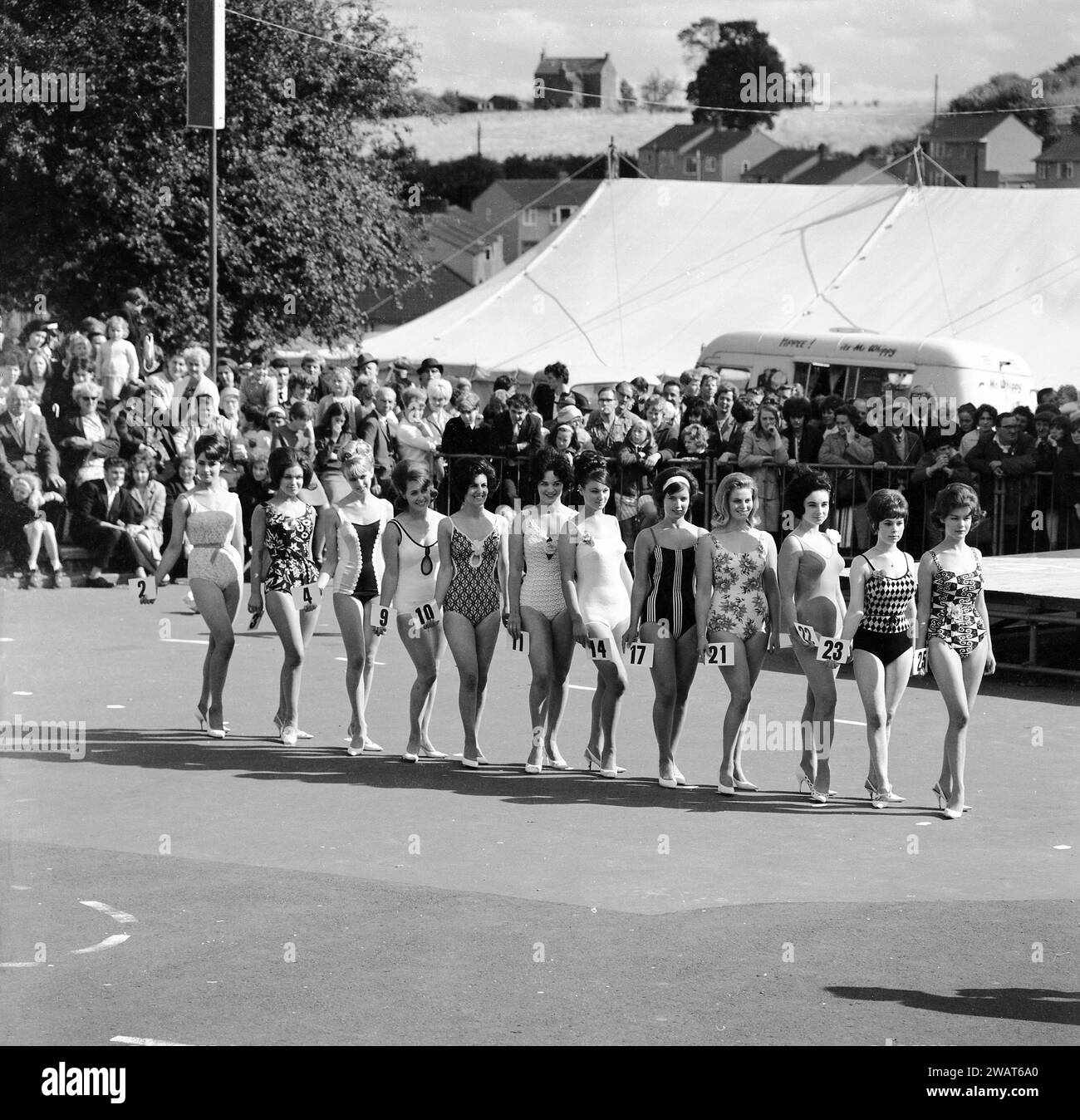 Années 1960, historiques, les spectateurs regardant un concours de reine de beauté, tandis que les dames en maillot de bain et tenant des numéros de concours, s'alignent pour une photo de groupe, Angleterre, Royaume-Uni. Derrière la foule, une grande tente et un Happee ! M. Whippy van vend de la crème glacée molle. Dans la Grande-Bretagne d'après-guerre, les concours de beauté étaient une forme de divertissement populaire pour les personnes en vacances dans les stations balnéaires. En 1945, Morecambe dans le Lancashire voit le premier concours Miss Grande-Bretagne avoir lieu. Banque D'Images