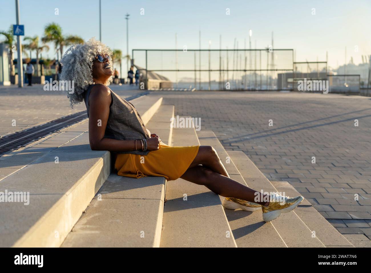Élégante dame afro-américaine avec des cheveux afro hoary et des lunettes de soleil se relaxant au soleil dans un environnement urbain. Concept : pro-vieillissement, vacances Banque D'Images