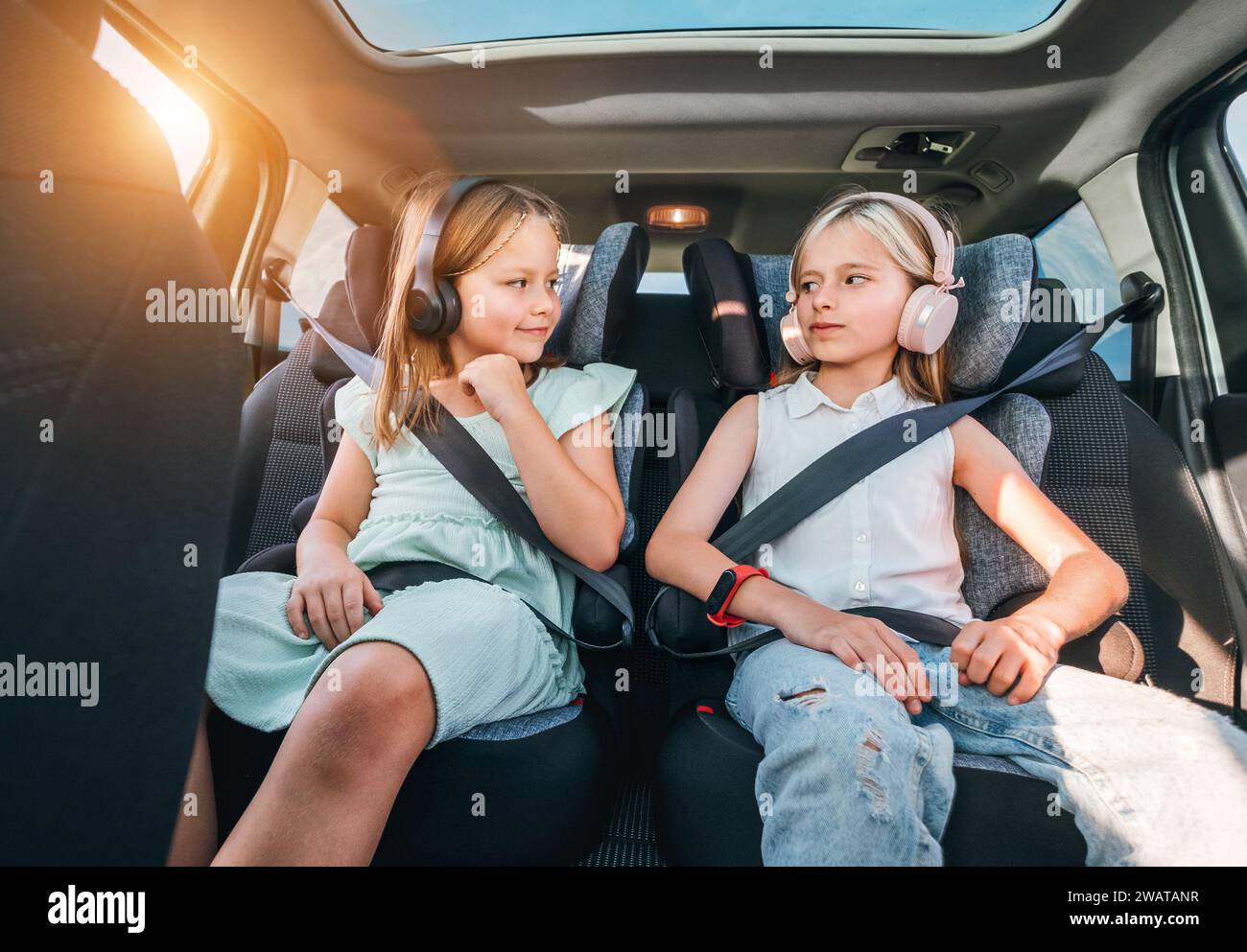 Portrait de deux sœurs souriantes positives assises dans des sièges d'auto pour enfants attachant des ceintures de sécurité en écoutant de la musique à l'aide d'écouteurs. Enfance heureuse, auto Banque D'Images