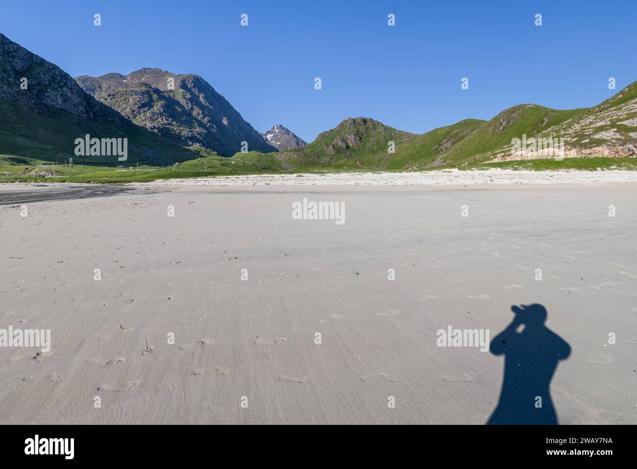 Une ombre du photographe s'étend sur le sable lisse de Haukland Beach, Lofoten, Norvège, avec des montagnes escarpées sous un ciel dégagé Banque D'Images