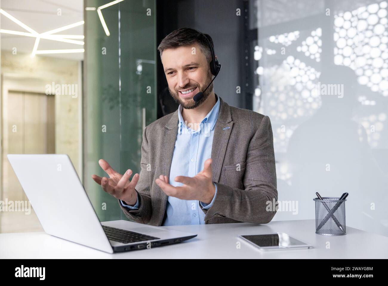Jeune homme souriant assis au bureau dans le bureau portant un casque et parlant en faisant des gestes mains lors d'un appel vidéo via un ordinateur portable. Banque D'Images