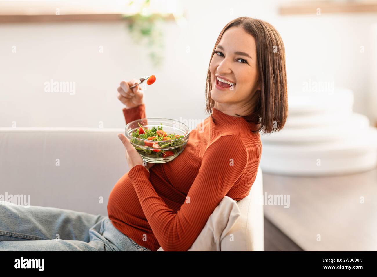 Heureuse femme enceinte mangeant salade assise sur le canapé à la maison Banque D'Images