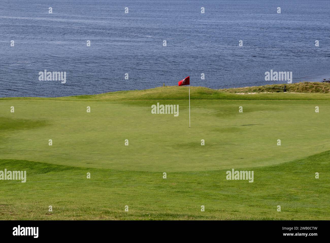 Un drapeau rouge et noir sur un bâton blanc dans un trou de golf le long de l'eau. L'herbe verte luxuriante est trempée et élevée avec son point le plus bas au milieu. Banque D'Images