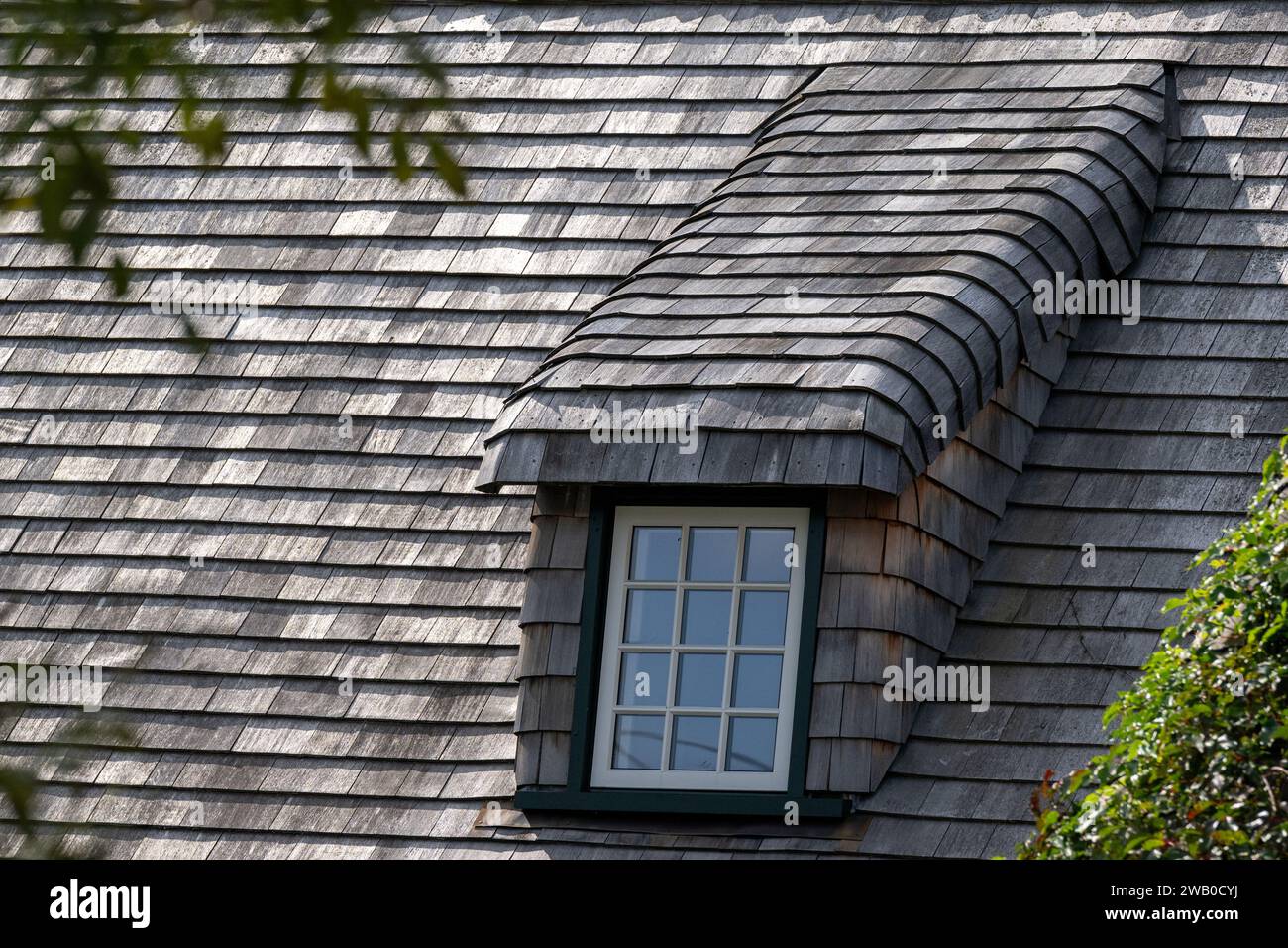 Un toit escarpé d'un bâtiment vintage avec des bardeaux en bois de cèdre. Il y a une longue lucarne recouverte de bois altéré. Le cadre de la fenêtre est de couleur verte. Banque D'Images