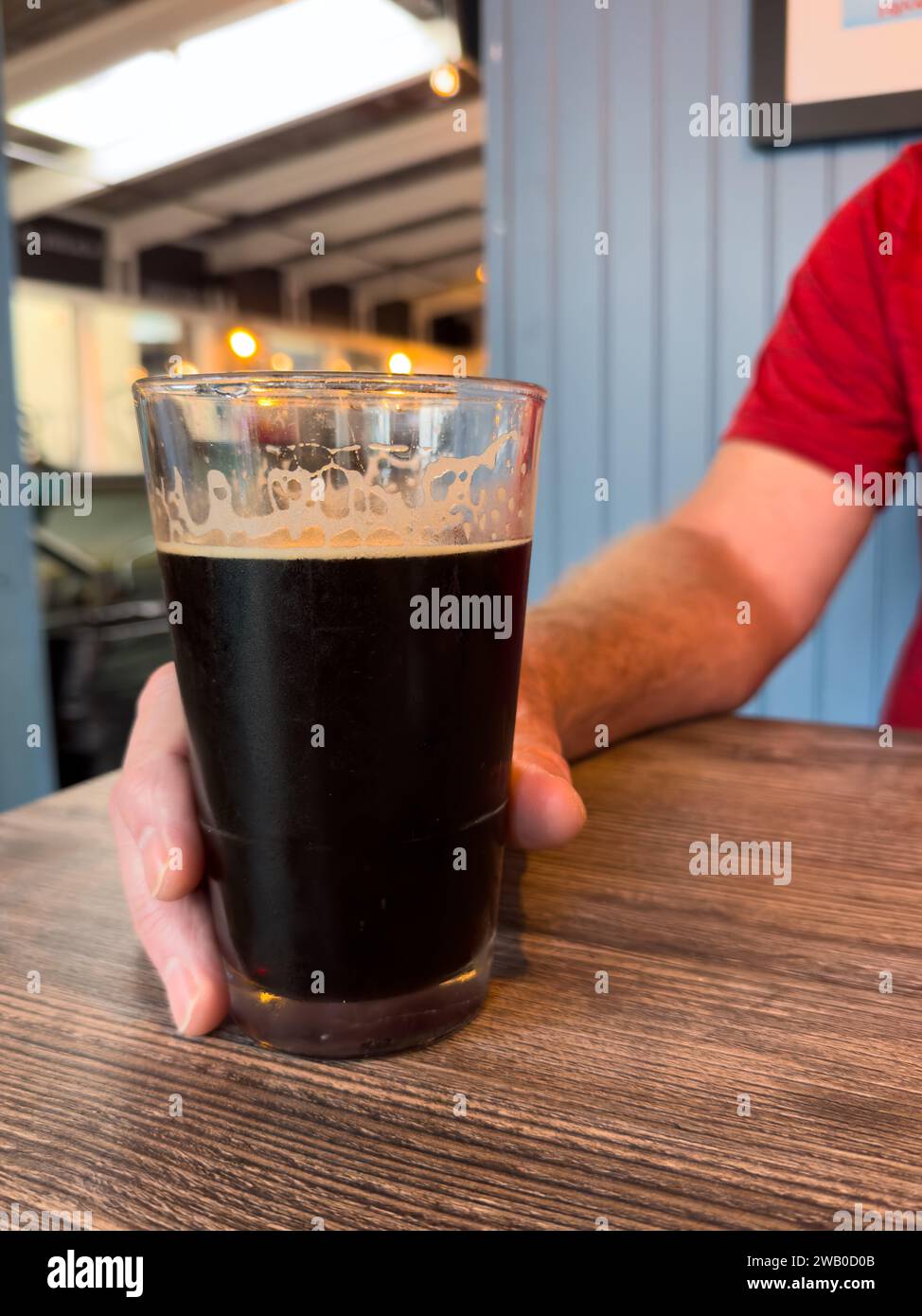 Un homme portant un t-shirt rouge vif tient une pinte de bière brune sur une table en bois. Le porteur de chocolat amer froid a de la mousse et des bulles. Banque D'Images