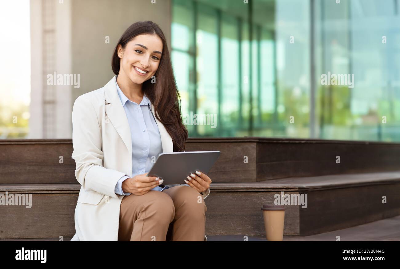 Joyeuse jeune femme professionnelle assise sur un banc avec une tablette numérique, smartwatch sur son poignet Banque D'Images