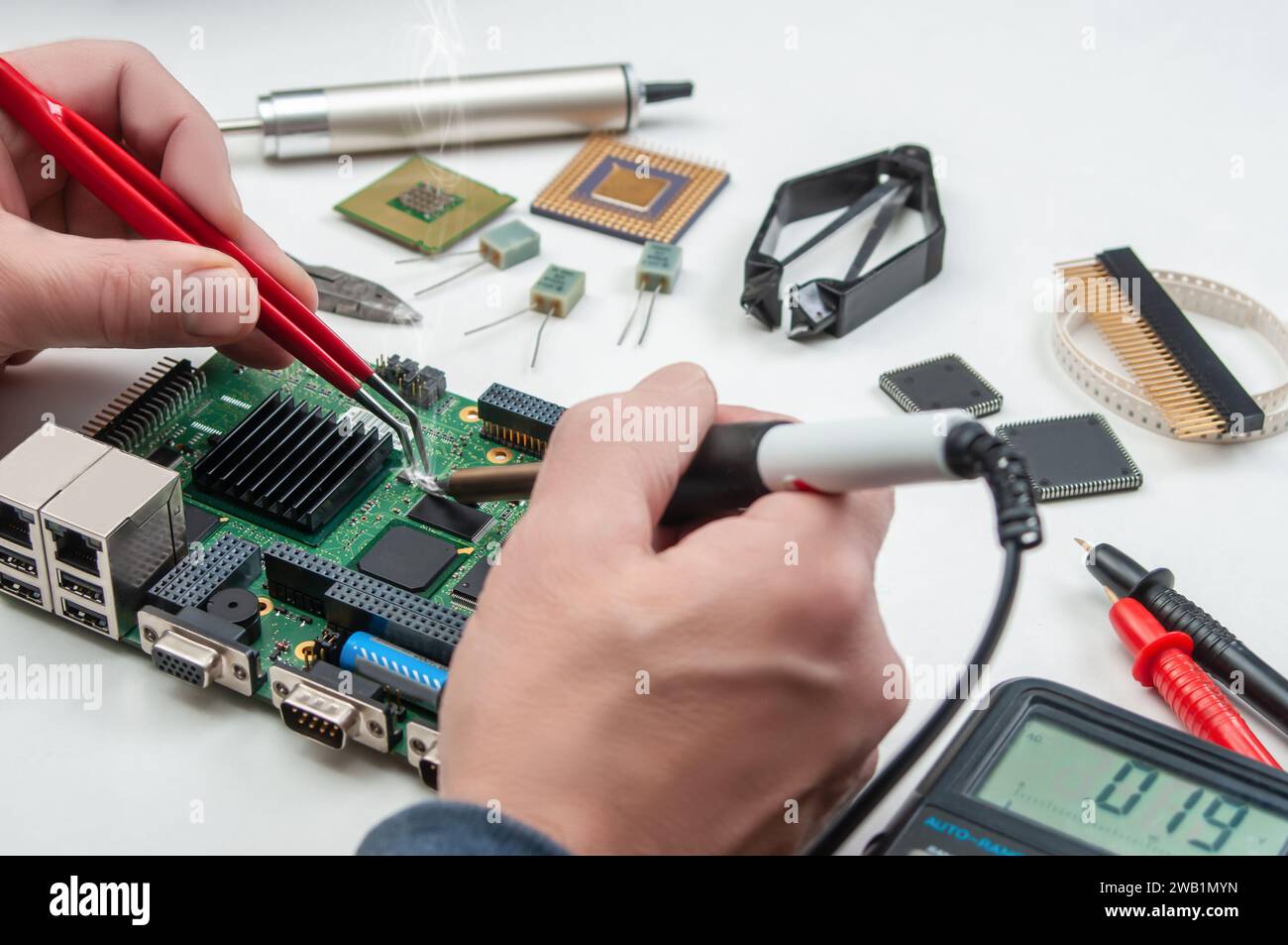 Gros plan des mains d'un technicien dans un atelier. Réparateur soude la carte de circuit imprimé de l'appareil électronique sur la table avec des brucelles. Banque D'Images