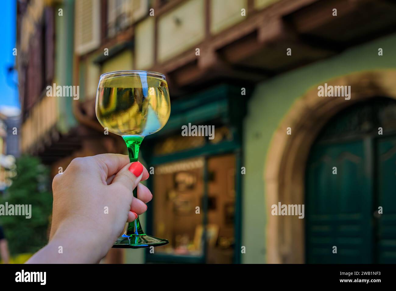 Main de femme avec un verre de vin Gewurztraminer dans un restaurant en plein air, maisons à colombages floues à Colmar France, route des vins d'Alsace village Banque D'Images