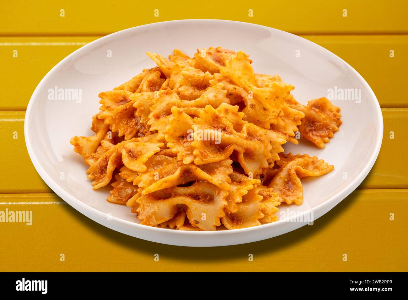 Pâtes Farfalle avec sauce tomate et Chili et ail et huile d'olive dans une assiette blanche isolée sur table en bois jaune avec chemin de détourage inclus Banque D'Images