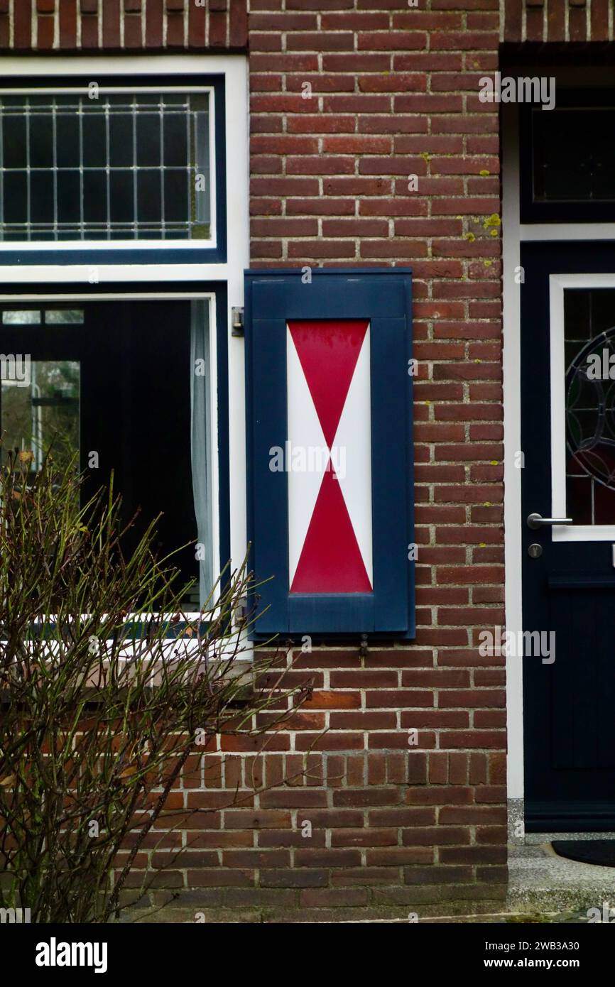 Un volet en bois bleu avec une croix diagonale rouge monté sur un mur de briques à côté d'une fenêtre et d'une porte. Banque D'Images