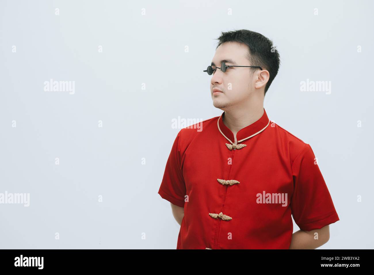 Visage inexpressif de l'homme asiatique portant Cheongsam et lunettes de soleil vintage noir tout en regardant le côté gauche sur fond blanc Banque D'Images