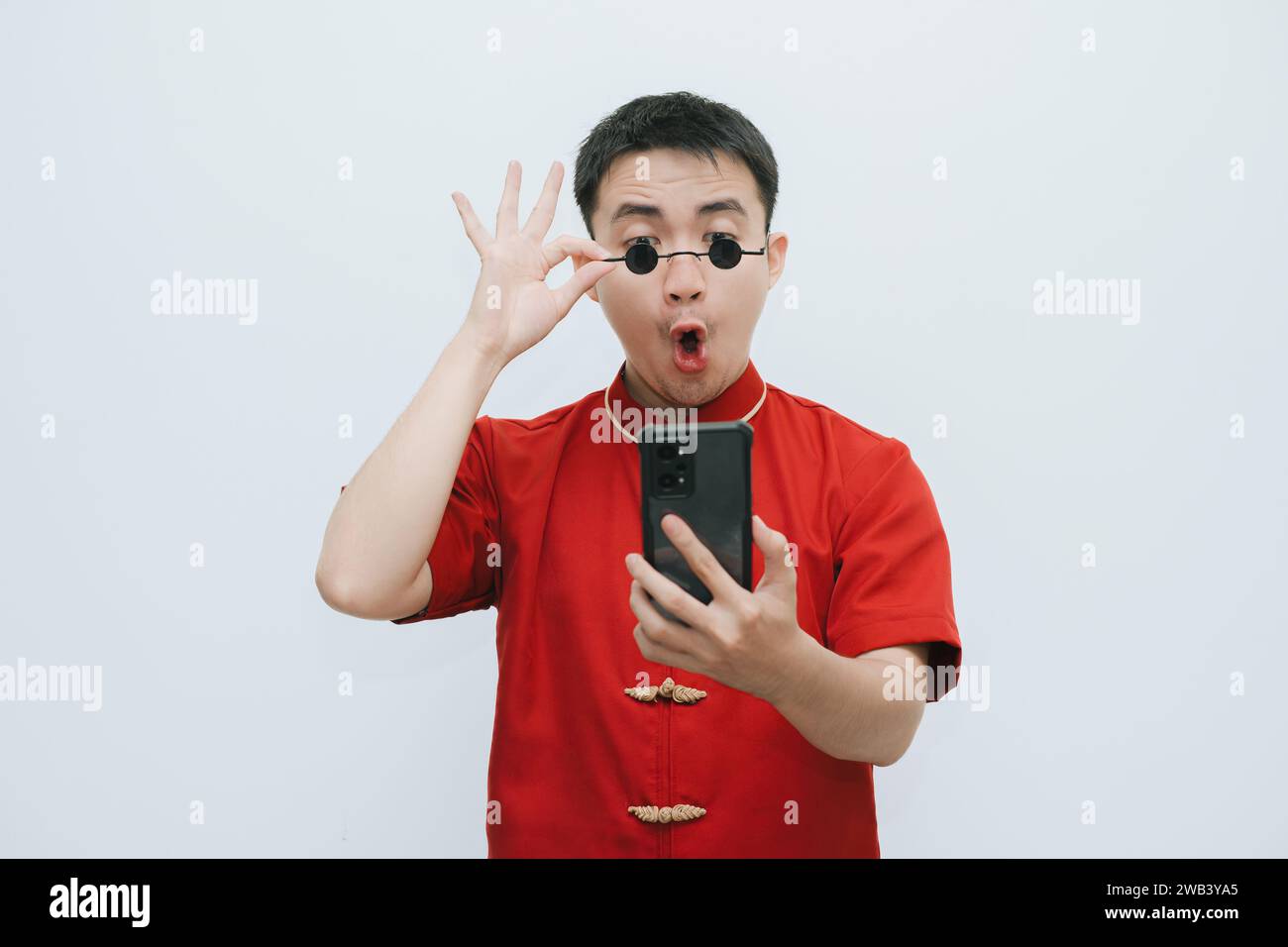 Visage choqué de l'homme asiatique portant Cheongsam et lunettes de soleil vintage noir tout en regardant le smartphone sur fond blanc Banque D'Images