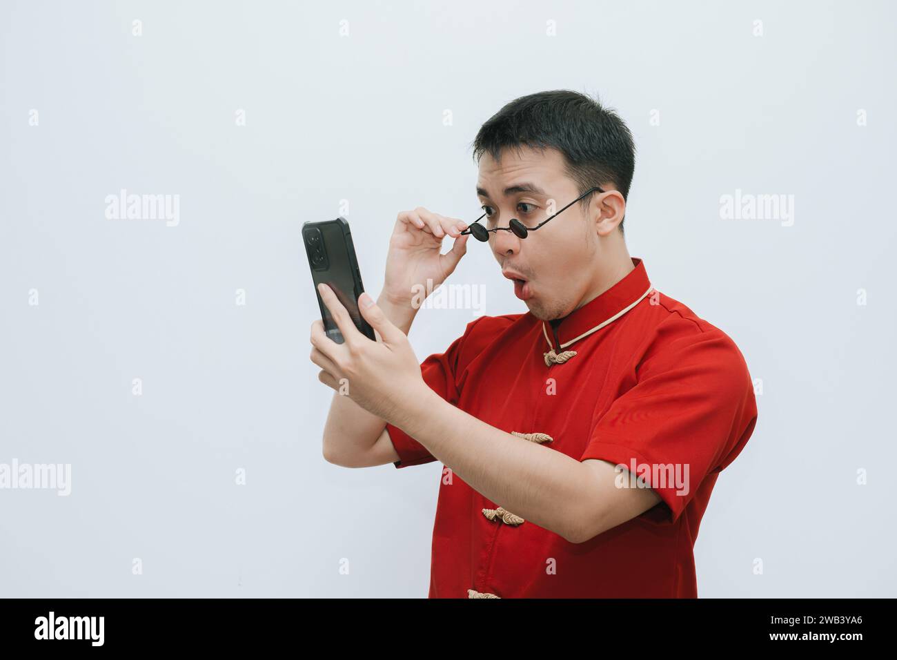 Visage choqué de l'homme asiatique portant Cheongsam et lunettes de soleil vintage noir tout en regardant le smartphone sur fond blanc Banque D'Images