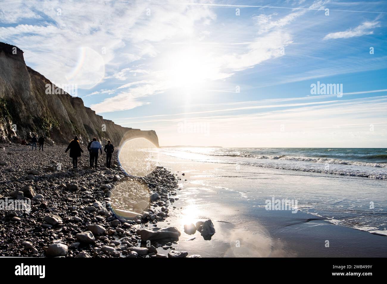 La photographie représente un groupe de personnes marchant le long d'une étendue rocheuse d'une plage avec d'imposantes falaises d'un côté. Le soleil du matin brille, créant un effet d'éclat et projetant les ombres des marcheurs sur le rivage de galets. Les vagues rencontrent doucement la plage, contribuant à l'atmosphère sereine du matin. Les falaises captent la lumière, mettant en valeur leurs textures et leurs couches, qui parlent de l’histoire géologique du littoral. L'image capture la tranquillité d'une promenade matinale au bord de la mer et l'interaction intemporelle entre la terre, la mer et le ciel. Promenade côtière sous le soleil du matin Elevé Banque D'Images